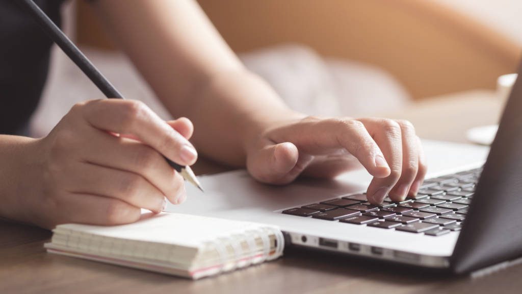 Close-up of student's hands, one hand holding a pencil and the other typing on a laptop keyboard